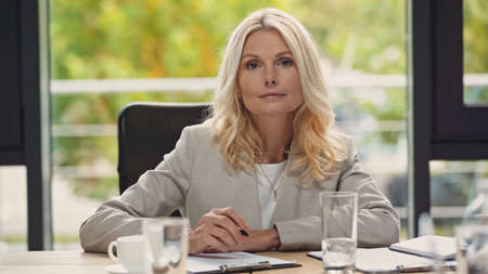 blonde middle aged businesswoman holding pen and looking at camera in meeting roomの写真素材