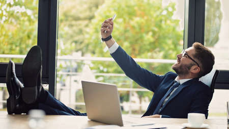 cheerful businessman taking selfie while sitting with legs on desk at workplaceの写真素材