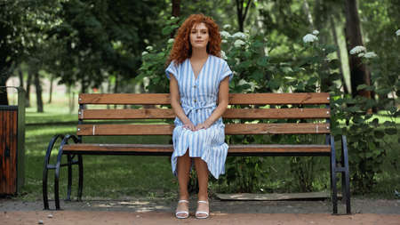full length of redhead woman in blue striped dress sitting on bench in park