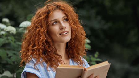 dreamy redhead woman smiling while holding book outdoorsの写真素材