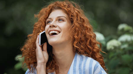 happy young woman with red hair laughing while talking on smartphoneの写真素材