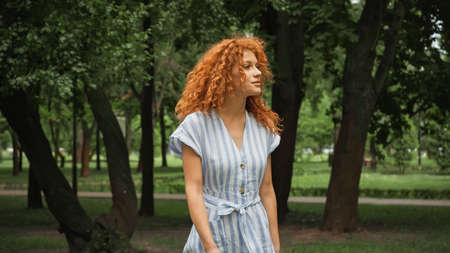 pretty young woman with red hair standing in blue striped dress in green park
