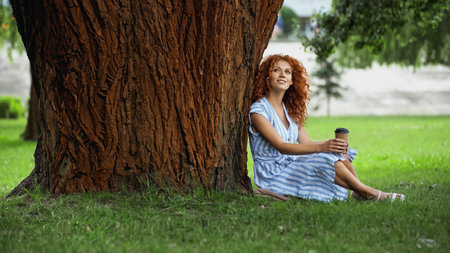 curly redhead woman in blue striped dress sitting under tree trunk with paper cupの写真素材