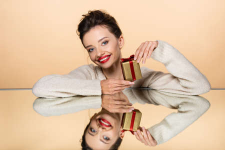 Smiling woman with red lips holding present near mirror isolated on beige