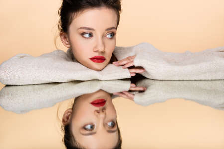 Pretty model with red lips wearing sweater and looking away near mirror isolated on beigeの写真素材