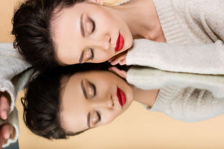 Stylish model in sweater closing eyes while lying on mirror isolated on beige