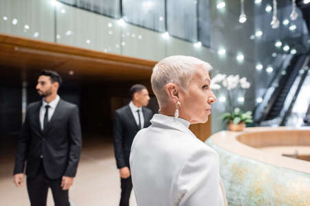 mature and stylish businesswoman looking away near interracial bodyguards in modern hotelの写真素材