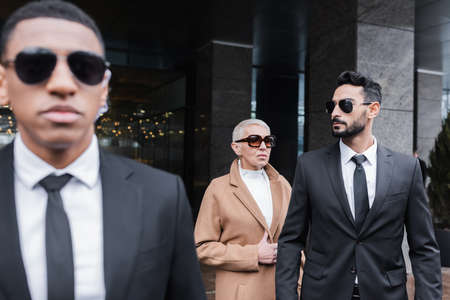 multiethnic security men in sunglasses protecting mature business lady near hotel building