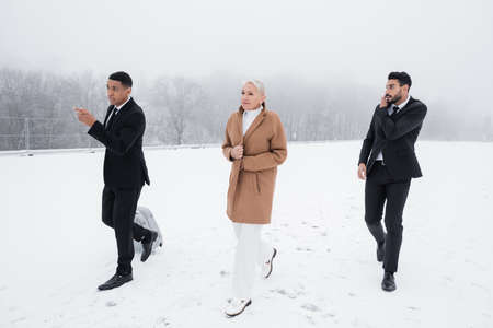 african american bodyguard with suitcase pointing direction near senior businesswoman on snowy field