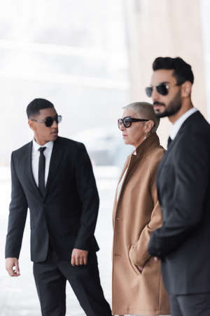 selective focus of wealthy businesswoman with hand in pocket near private interracial bodyguards...