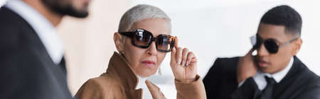 stylish business lady adjusting sunglasses near blurred multiethnic bodyguards outdoors, banner