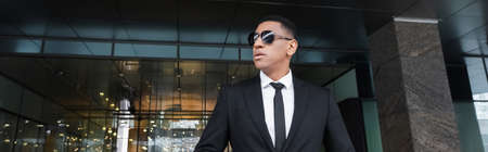 african american security man in formal wear and black sunglasses looking away near hotel entrance,...
