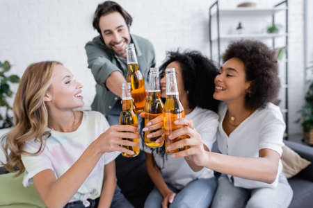 blurred happy man and joyful interracial women toasting bottles of beer