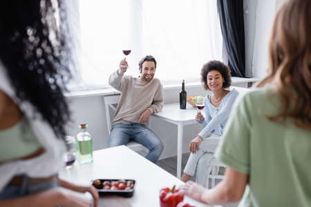 cheerful multiethnic friends holding glasses of red wine near blurred women in modern kitchen