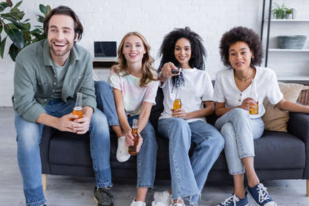 cheerful african american woman clicking channels near happy friends sitting on couch and holding beer in living roomの写真素材