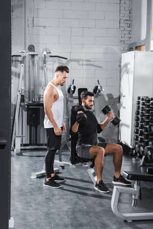 Young trainer standing near arabian sportsman training with dumbbells in gym