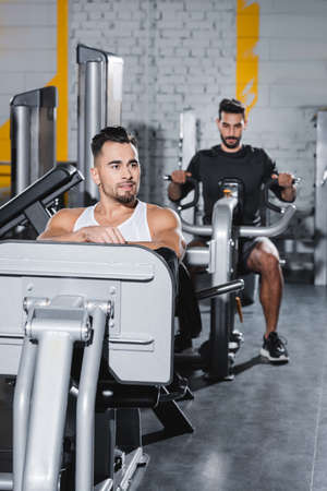 Smiling sportsman training on leg press machine near blurred arabian friend in gym