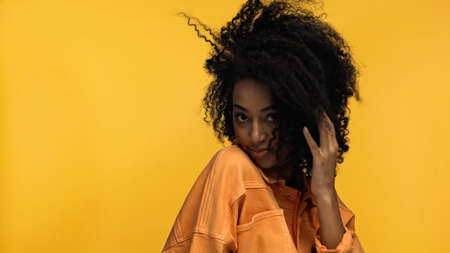 young and pleased african american woman adjusting curly hair isolated on yellow