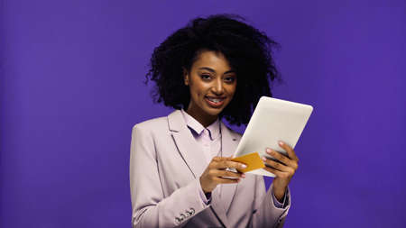 cheerful african american woman in blazer holding digital tablet and credit card isolated on purpleの写真素材