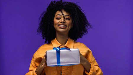 cheerful african american woman with braces holding wrapped present isolated on purpleの写真素材