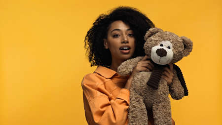 young african american woman with braces holding soft toy isolated on yellowの写真素材