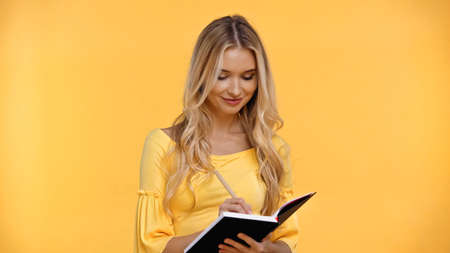 Smiling blonde woman writing on notebook isolated on yellowの写真素材
