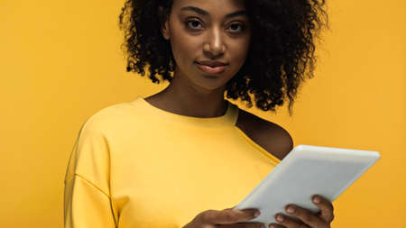 curly african american woman holding digital tablet isolated on yellowの写真素材