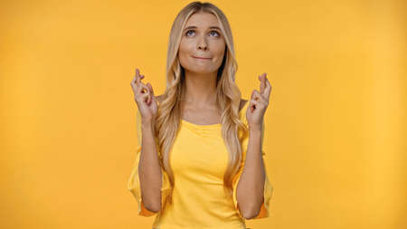 Blonde woman crossing fingers and looking up isolated on yellow
