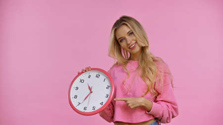 Smiling blonde woman pointing with finger at clock isolated on pink