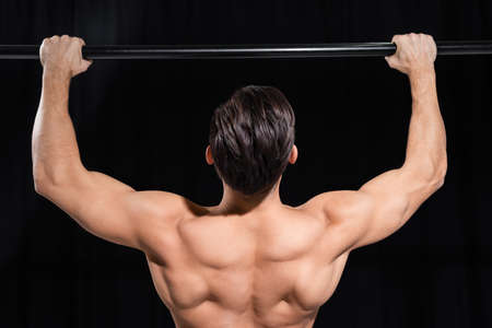 back view of muscular sportsman exercising on horizontal bar isolated on black
