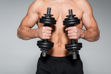 partial view of shirtless and muscular sportsman working out with dumbbells isolated on grayの写真素材
