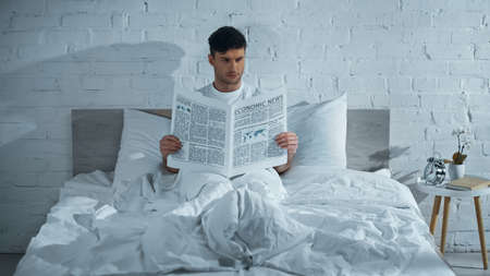 man reading newspaper while sitting on bed under white blanketの写真素材