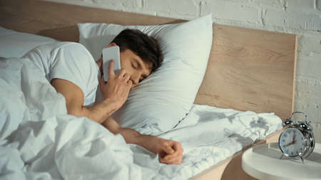 man with closed eyes talking on mobile phone in bed near alarm clock on bedside table