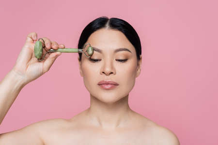 Asian woman with naked shoulders massaging eyebrow with jade roller isolated on pinkの写真素材