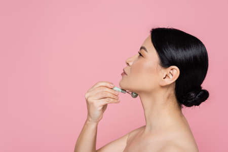 Side view of asian woman with naked shoulders massaging neck with jade roller isolated on pinkの写真素材