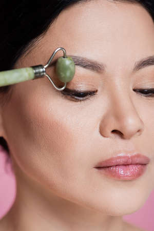 Close up view of asian woman massaging face with blurred jade roller isolated on pinkの写真素材