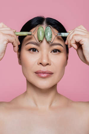 Asian woman with naked shoulders massaging forehead with jade rollers isolated on pinkの写真素材
