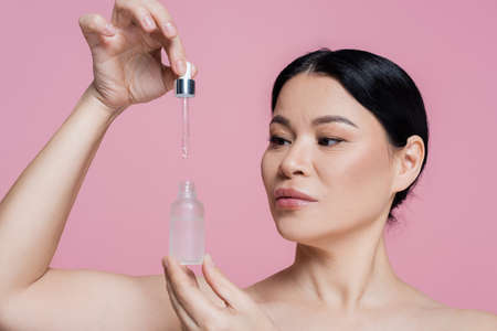 Asian woman with naked shoulders holding dropper and serum isolated on pinkの写真素材