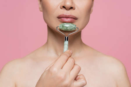 Cropped view of woman with naked shoulders massaging chin with jade roller isolated on pinkの写真素材