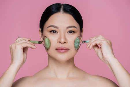 Asian woman with naked shoulders massaging cheeks with jade rollers isolated on pinkの写真素材