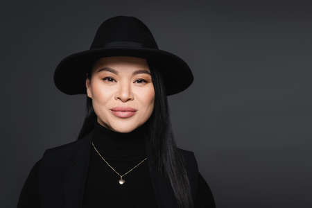 Portrait of stylish asian woman in fedora hat looking at camera isolated on dark grayの写真素材