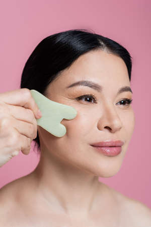 Asian woman massaging cheek with jade stone isolated on pinkの写真素材