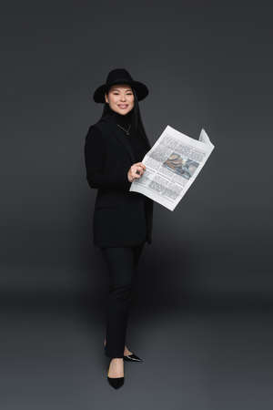 Full length of smiling asian model in fedora hat holding newspaper on dark gray backgroundの写真素材