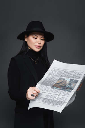 Asian woman in fedora hat and blazer reading newspaper isolated on dark grayの写真素材