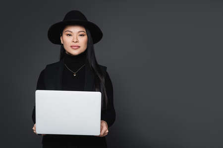 Stylish asian woman in fedora hat and turtleneck holding laptop isolated on dark grayの写真素材