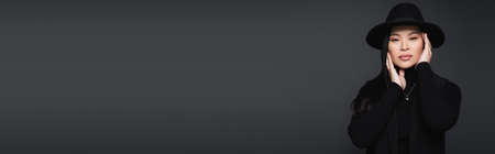 Trendy asian woman in fedora hat touching face isolated on dark grey, bannerの写真素材