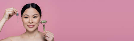 Asian woman with naked shoulders holding jade roller and serum isolated on pink, bannerの写真素材