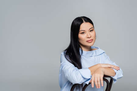 Asian woman in blouse looking at camera near chair isolated on grayの写真素材