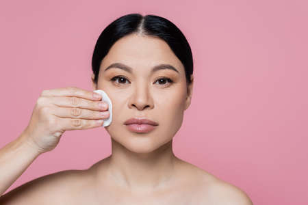 Asian woman with naked shoulders cleaning face with cotton pad isolated on pinkの写真素材