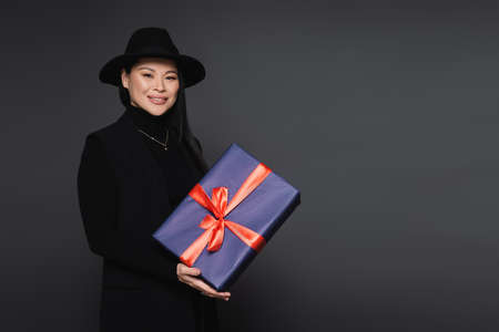 Smiling asian woman in fedora hat holding present isolated on dark grayの写真素材